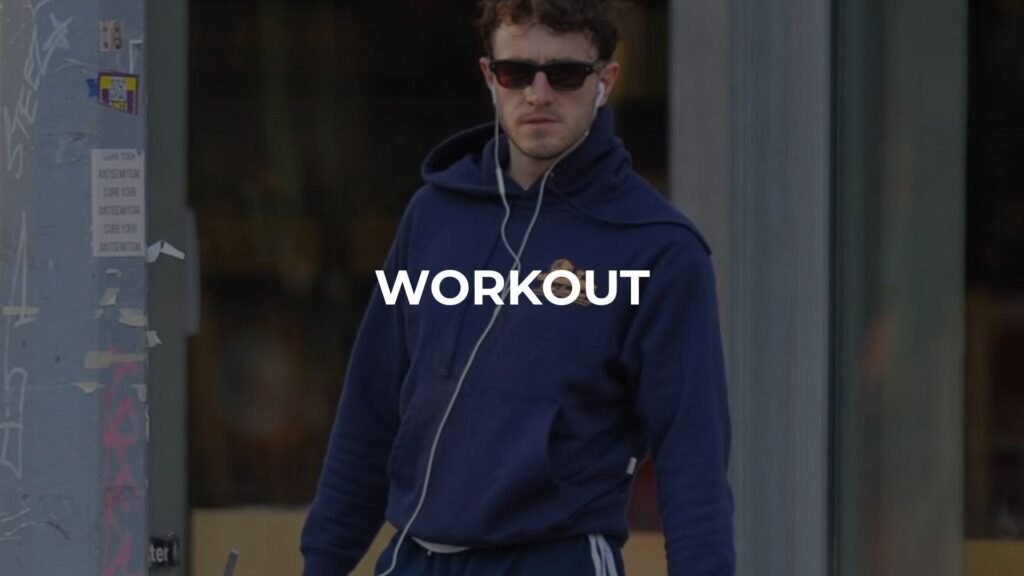 Hombre joven con cabello rizado, lentes de sol, sudadera azul marino con capucha, pants y audífonos con cable, caminando por una calle de la ciudad con la palabra "WORKOUT" superpuesta en el centro.