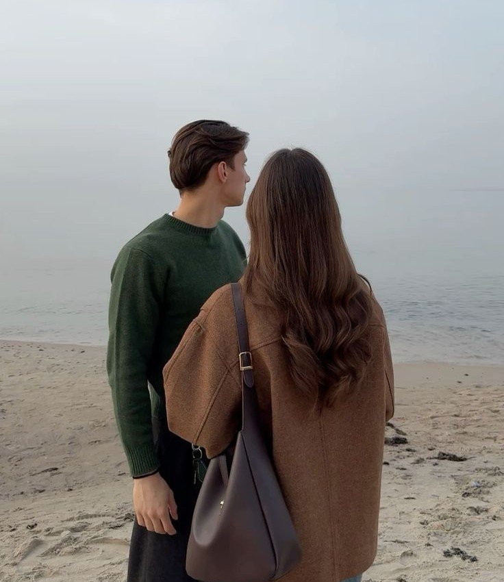 Pareja de espaldas en la playa con look casual de otoño, cabello largo con ondas y ambiente nublado en la playa.